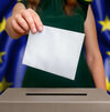 Hand wirft Wahlumschlag in Wahlurne vor Europaflagge